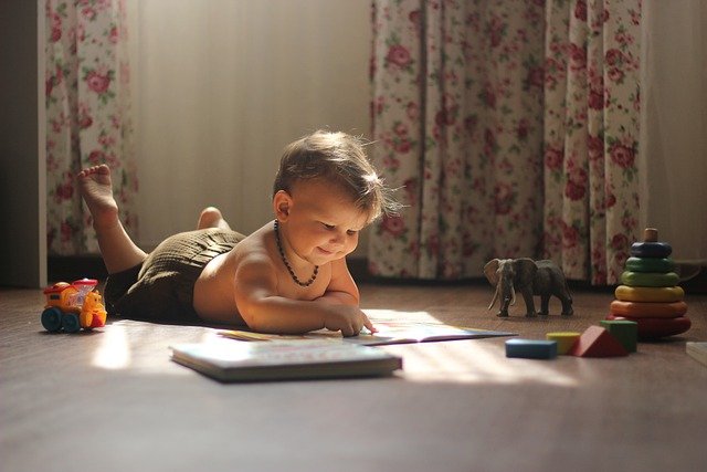Baby reading book on the floor surrounded by toys