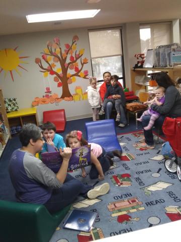 Librarian reading books to children and parents on colorful "READ" rug